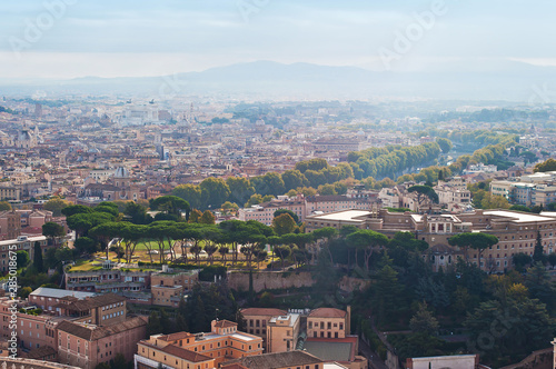 View of many orange rooftops and rows of high green trees and outline of a smoky mountain range, Rome