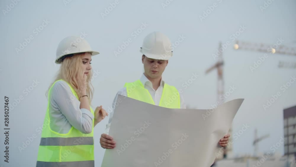 Two engineers discuss looking at the drawings of the construction plan and the location of the objects specifying the contractor on the phone details. Talking on the phone at the construction site Two engineers discuss looking at the drawings of the construction plan and the location of the objects specifying the contractor on the phone details. Talking on the phone at the construction site