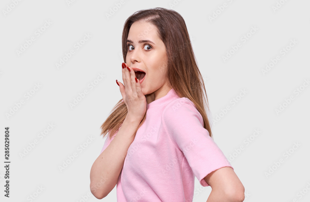Shoked young woman has amazed expression, wearing pink shirt. Portrait of pretty amazed female, holding hand on mouth with red nails, looking astonished posing over white background. People emotions Shoked young woman has amazed expression, wearing pink shirt. Portrait of pretty amazed female, holding hand on mouth with red nails, looking astonished posing over white background. People emotions