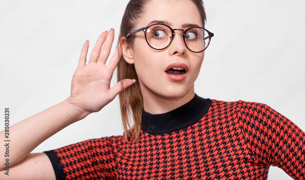 Amazed young woman paying attention and placing hand on ear asking someone to speak louder or whisper, isolated on white wall. Pretty girl which overhears secret conversation, wearing eyeglasses. Amazed young woman paying attention and placing hand on ear asking someone to speak louder or whisper, isolated on white wall. Pretty girl which overhears secret conversation, wearing eyeglasses.