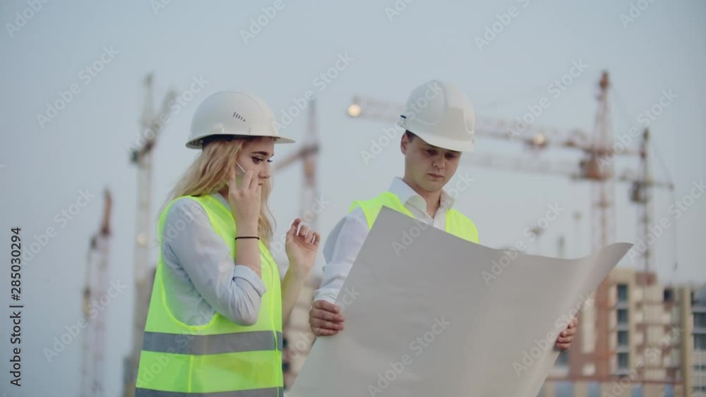 Woman talking on the phone and asks the Builder what is on the drawings standing on the background of buildings under construction Woman talking on the phone and asks the Builder what is on the drawings standing on the background of buildings under construction