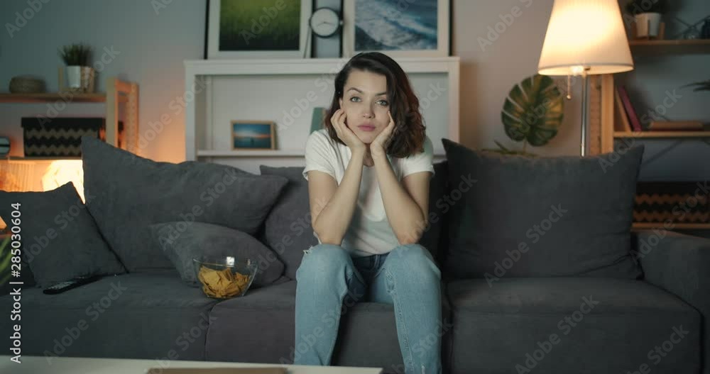 Young beautiful woman is watching TV with serious face at home sitting on couch alone enjoying domestic entertainment at night. People, television and apartment concept. Young beautiful woman is watching TV with serious face at home sitting on couch alone enjoying domestic entertainment at night. People, television and apartment concept.