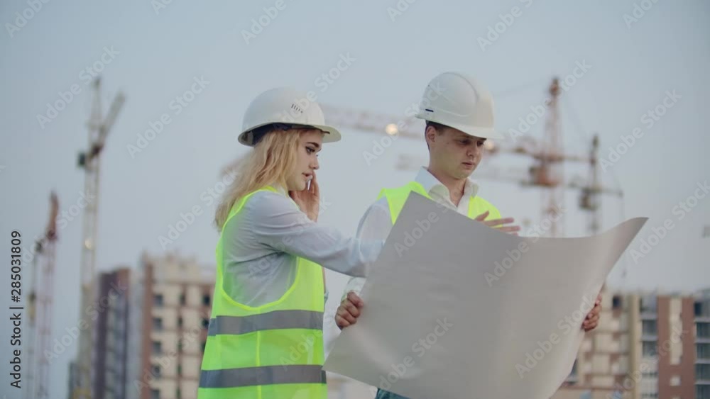 Two engineers discuss looking at the drawings of the construction plan and the location of the objects specifying the contractor on the phone details. Talking on the phone at the construction site Two engineers discuss looking at the drawings of the construction plan and the location of the objects specifying the contractor on the phone details. Talking on the phone at the construction site