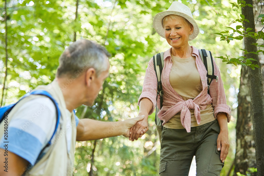 Senior man helping moving down the mature smiling woman while they have a trip in the forest in summer Senior man helping moving down the mature smiling woman while they have a trip in the forest in summer