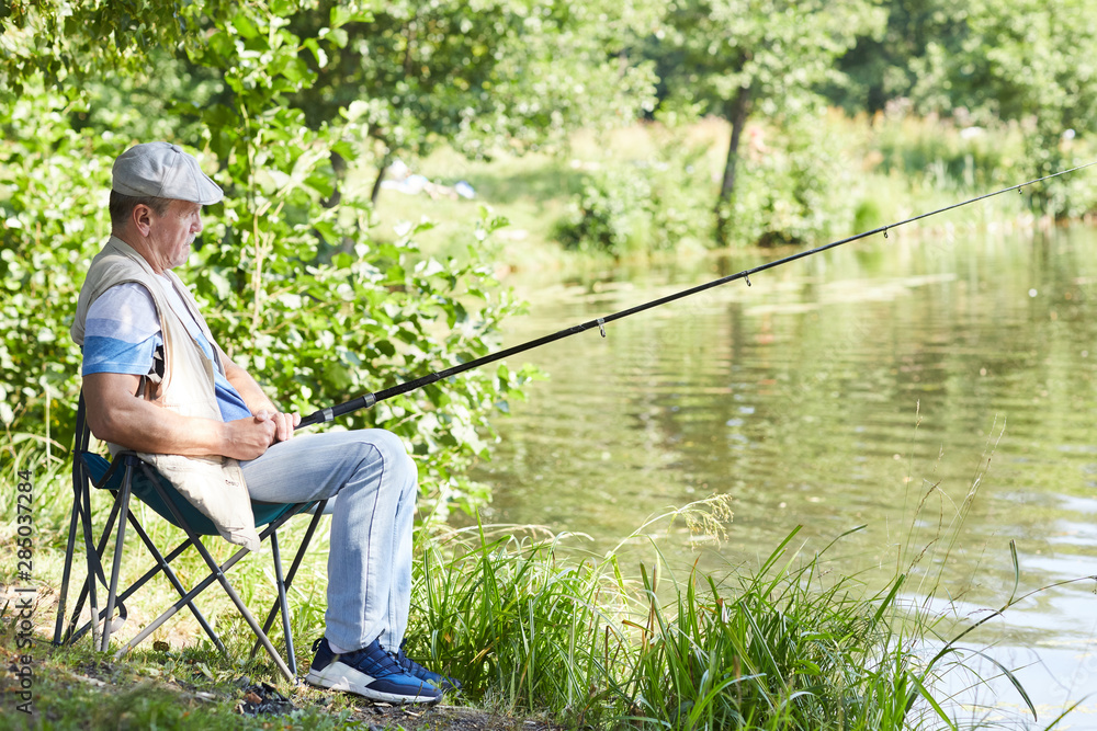 Senior man with fishing rod catching fish while sitting on chair near the lake outdoors Senior man with fishing rod catching fish while sitting on chair near the lake outdoors