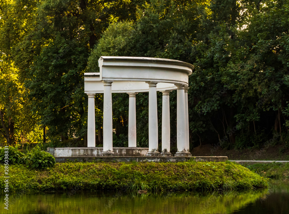 A small building with white columns on the greenery of the park