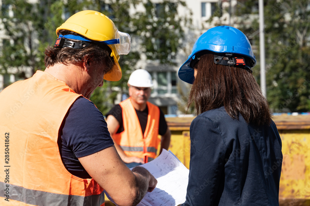 Construction manager wearing safety jacket and helmet checking projects discussing with a female engineer. In background, another worker Construction manager wearing safety jacket and helmet checking projects discussing with a female engineer. In background, another worker