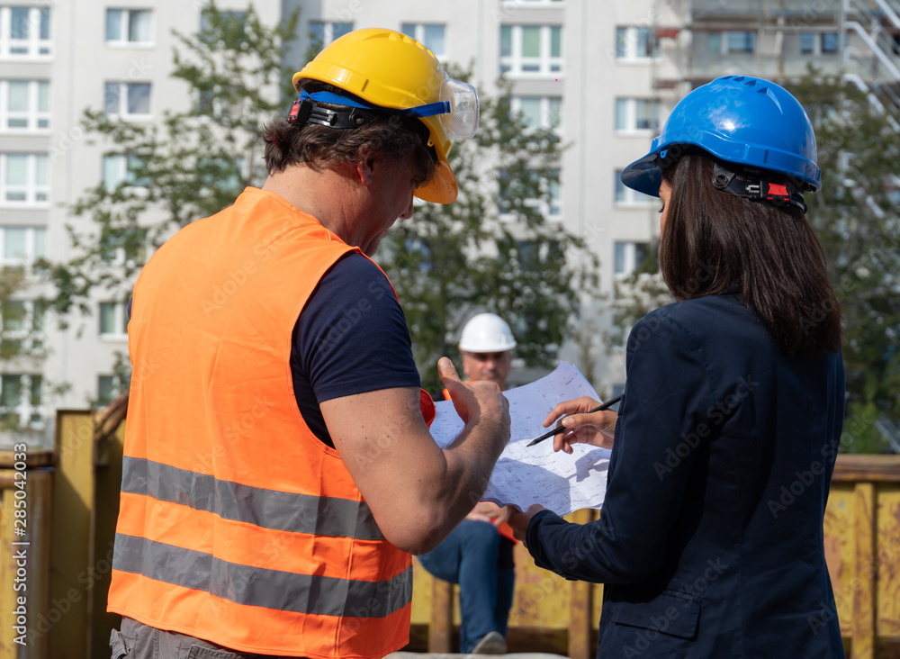 Construction manager wearing safety jacket and helmet checking projects discussing with a female engineer. In background, another worker Construction manager wearing safety jacket and helmet checking projects discussing with a female engineer. In background, another worker