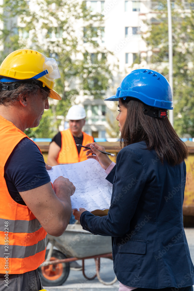 Construction manager wearing safety jacket and helmet checking projects discussing with a female engineer. In background, another worker Construction manager wearing safety jacket and helmet checking projects discussing with a female engineer. In background, another worker