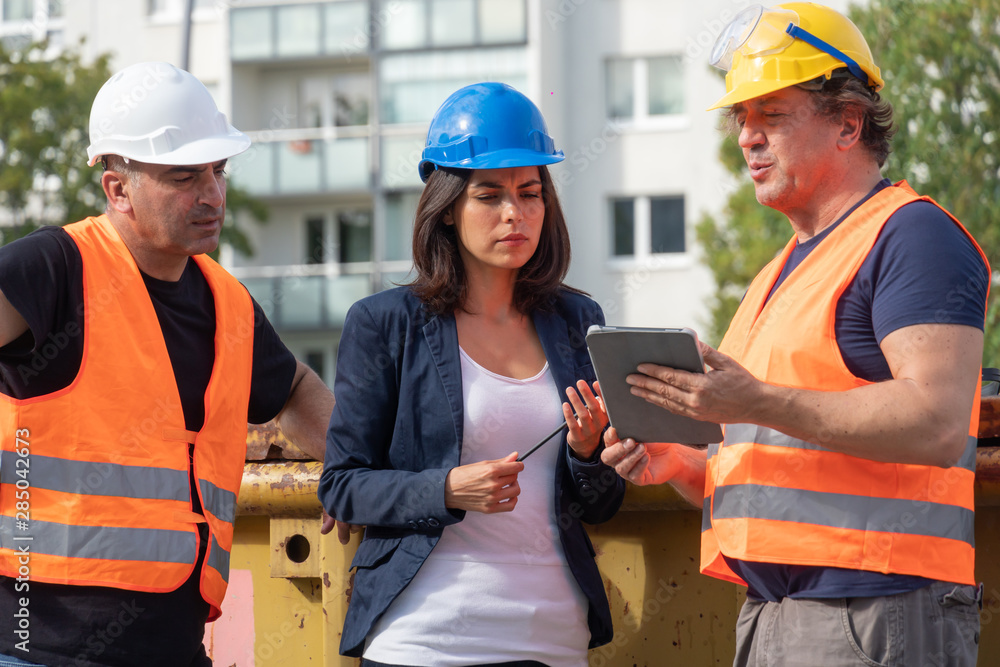 Senior foreman wearing safety vest, helmet and goggles providing instructions to a male and a female colleagues using a digital tablet computer on construction site Senior foreman wearing safety vest, helmet and goggles providing instructions to a male and a female colleagues using a digital tablet computer on construction site