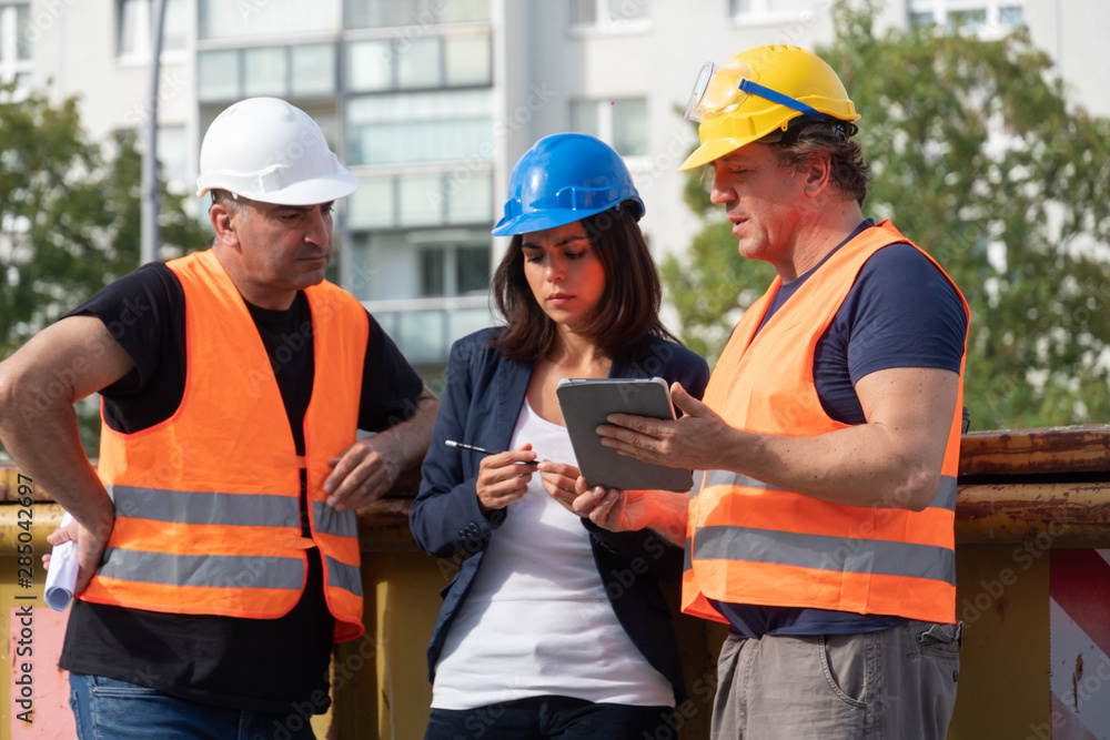 Senior foreman wearing safety vest, helmet and goggles providing instructions to a male and a female colleagues using a digital tablet computer on construction site Senior foreman wearing safety vest, helmet and goggles providing instructions to a male and a female colleagues using a digital tablet computer on construction site