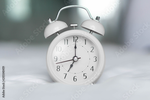 Alarm clock on white bad for wake up time with light from window, selective focus