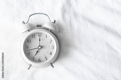 Alarm clock on white bad for wake up time with light from window, selective focus