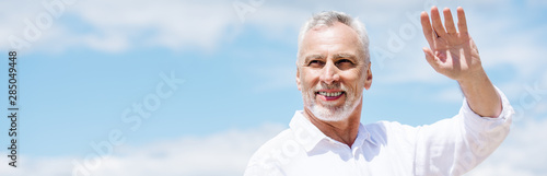 panoramic view of smiling senior man in white shirt waving hand under blue sky
