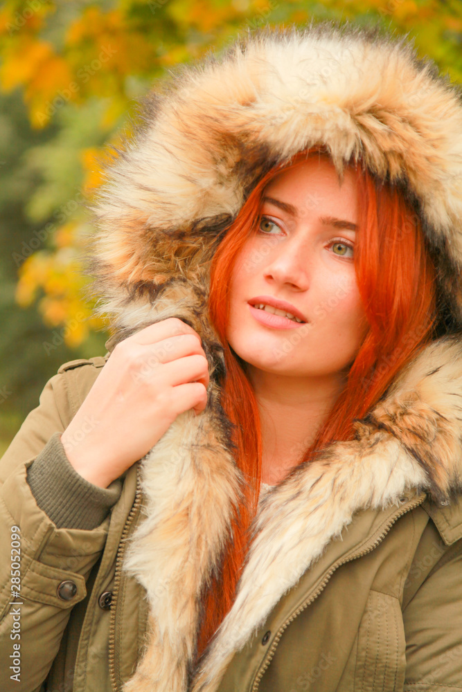 Redheaded woman in coat with fur hood in autumn park with yellow green trees Redheaded woman in coat with fur hood in autumn park with yellow green trees
