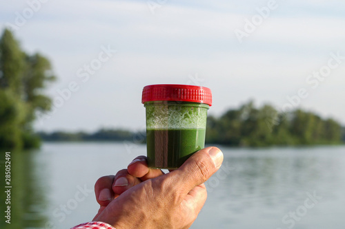 closeup hand holding container with green algae with blurred river on background