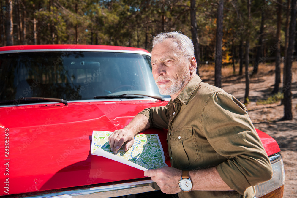 pensive senior tourist standing near red car and holding map in sunny day pensive senior tourist standing near red car and holding map in sunny day