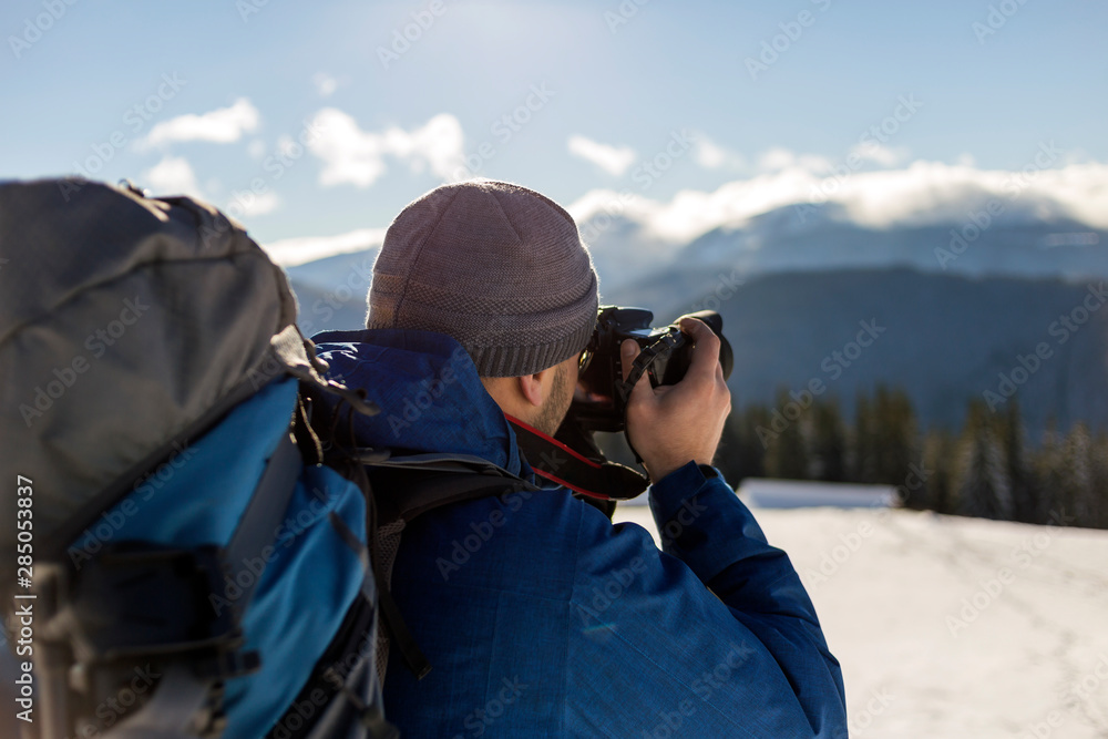 Hiker man tourist photographer in warm clothing with backpack and camera taking picture of snowy valley and woody mountain peaks landscape under blue sky on sunny winter cold day. Hiker man tourist photographer in warm clothing with backpack and camera taking picture of snowy valley and woody mountain peaks landscape under blue sky on sunny winter cold day.