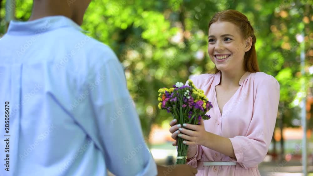 Mixed-race teen boy presenting flowers to surprised girlfriend, date in park Mixed-race teen boy presenting flowers to surprised girlfriend, date in park