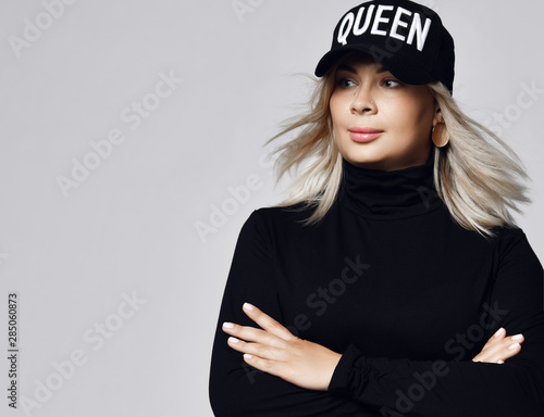 Portrait of woman with straight long blonde hair in black cap and sweater standing with hands crossed in front of her photo