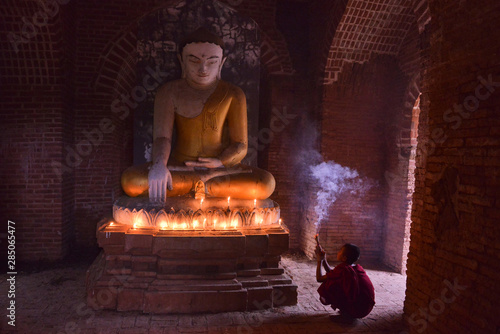 young budhist pilgrim  monk dressed in traditional red robe praying in front of buddha in a temple in myanmar