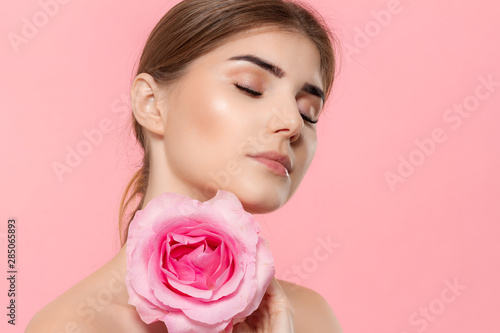 Close-up portrait of a beautiful young girl with pink rose flower isolated over pink background.