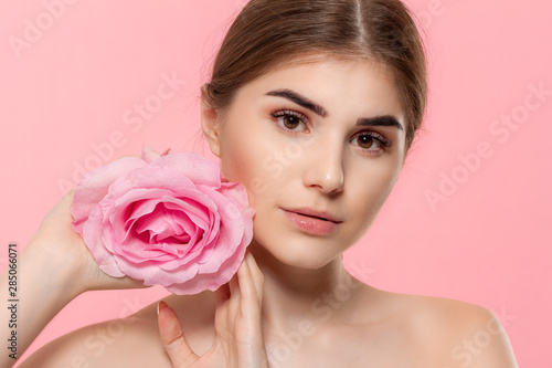 Close-up portrait of a beautiful young girl looking at the camera holding pink rose flower close to face isolated over pink background.