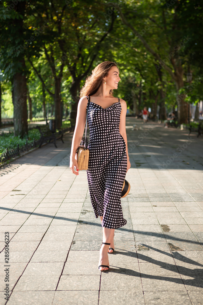 Beautiful young brunette woman dressed in a black dress and holding a hat with wide flaps takes a walk in a park during warm summer day enjoying sunlight.