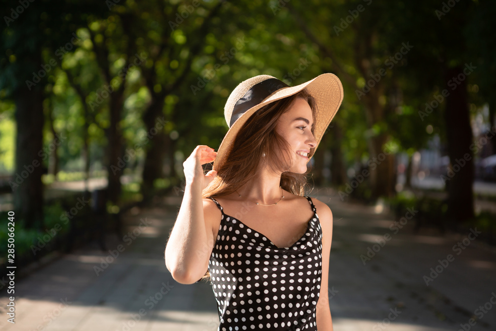 Beautiful young brunette woman dressed in a black dress and a hat with wide flaps takes a walk in a park during warm summer day enjoying sunlight.