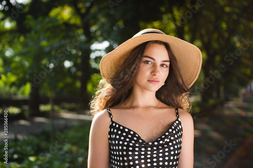 Close-up porttrait of a beautiful young brunette woman dressed in a black dress and a hat with wide flaps takes a walk in a park during warm summer day enjoying sunlight.