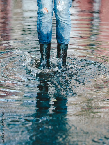 Legs of woman in shabby jeans with black rubber boots standing in a puddle of water.