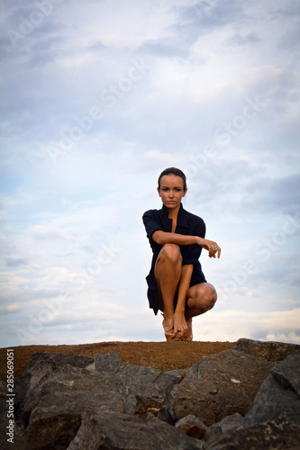 Fashion pretty young woman with long legs sitting outdoor on the ground near a Electric poles. Beautiful brunette on heels only in black shirt, bare feet