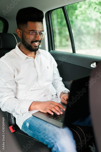 Full concentration at work. Confident indian young man in full suit working using laptop while sitting in the car