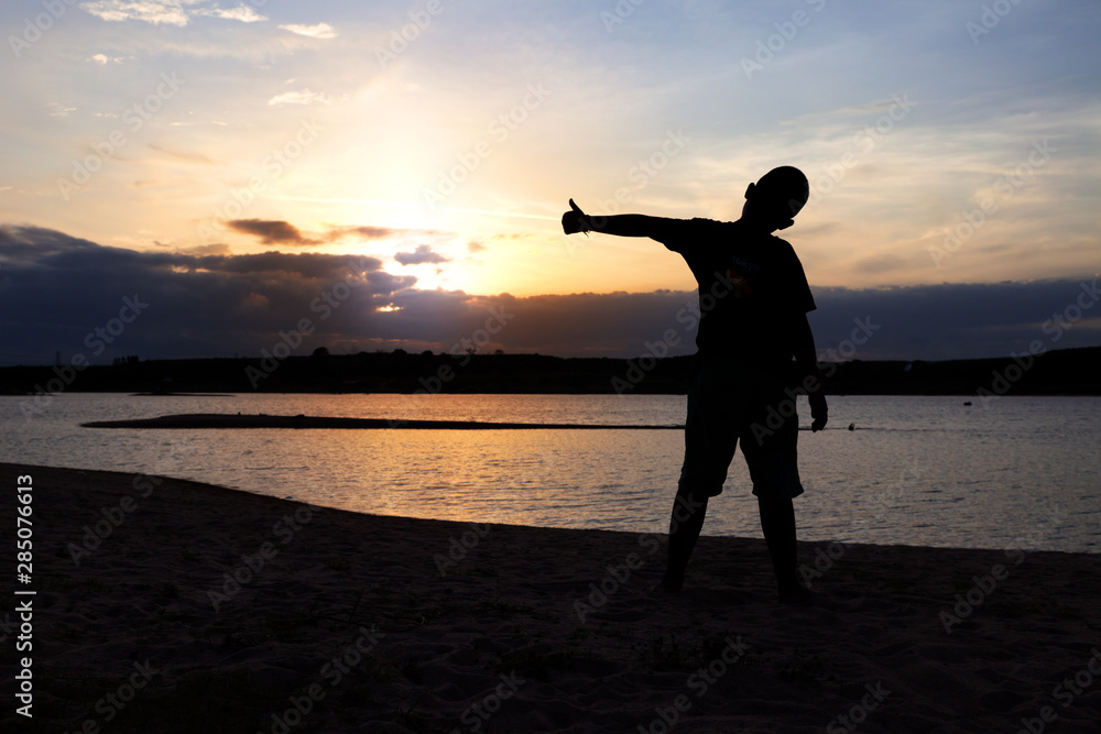 Fototapeta premium Silhouette of boy with arms spread out next to river sunset. Child on river sunset.