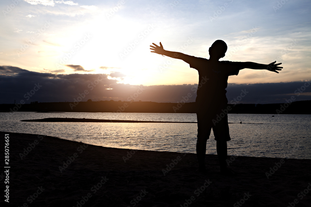 Fototapeta premium Silhouette of boy with arms spread out next to river sunset. Child on river sunset.