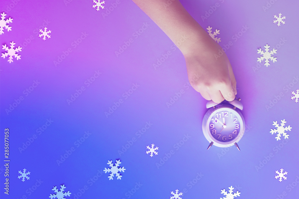 Child's hand holding alarm clock showing five to twelve with paper snowflakes, neon toning