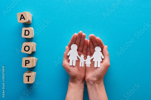 cropped view of woman holding paper cut lesbian family near blocks with adopt lettering on blue background