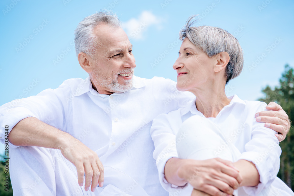 happy smiling senior couple in white shirts embracing and looking at each other under blue sky