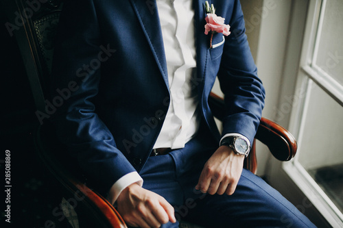 Stylish groom in blue suit, with bow tie and boutonniere with pink rose sitting in chair near window in hotel room. Morning preparation before wedding ceremony