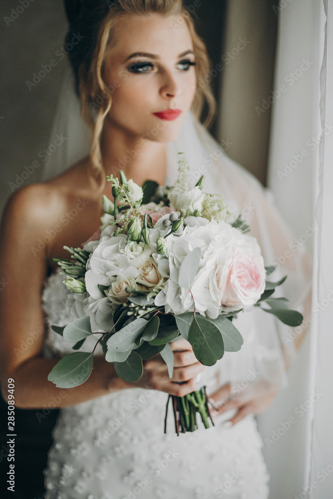 Fototapeta premium Stylish bride holding modern wedding bouquet and posing in soft light near window in hotel room. Gorgeous sensual bride portrait. Morning preparation before wedding ceremony