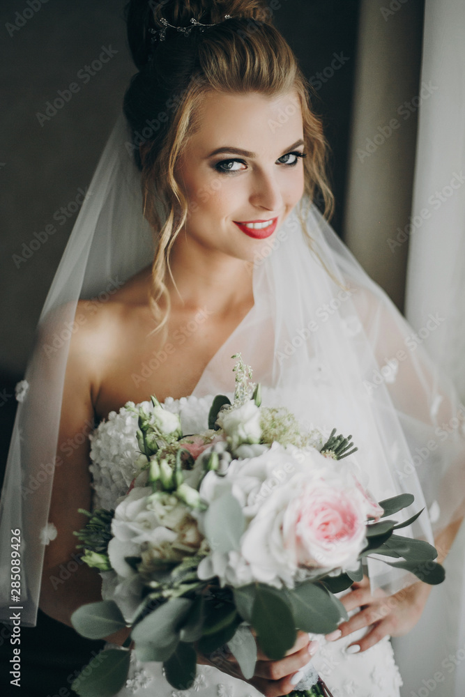 Naklejka premium Stylish bride holding modern wedding bouquet and posing in soft light near window in hotel room. Gorgeous sensual bride portrait. Morning preparation before wedding ceremony