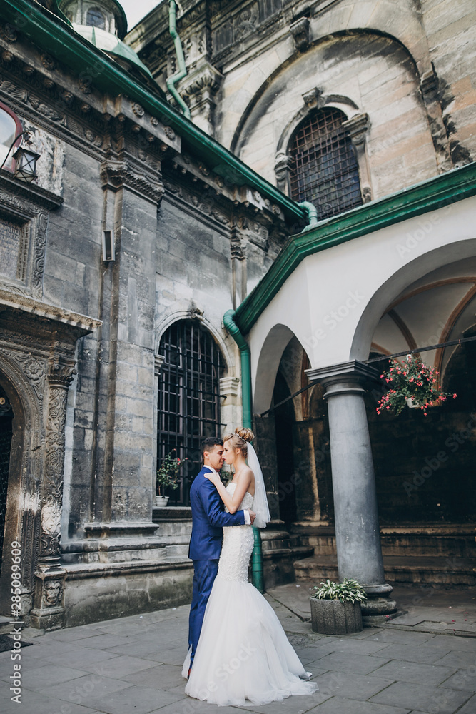 Stylish bride and groom gently hugging in sunny european city street. Gorgeous wedding couple of newlyweds embracing in old buildings. Romantic moment