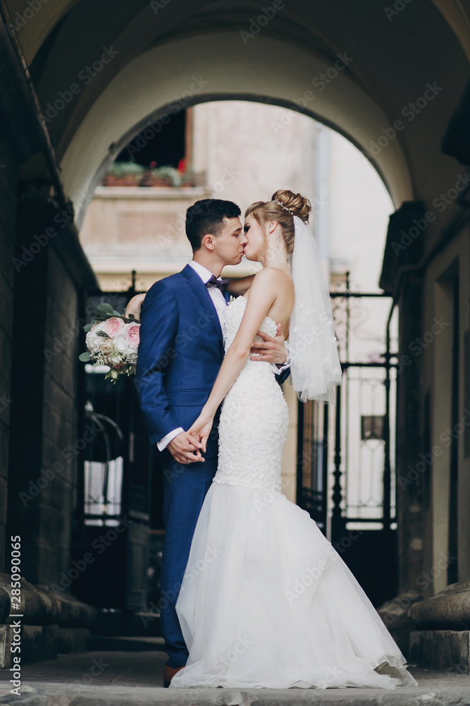 Stylish bride and groom gently kissing in sunny european city street. Gorgeous wedding couple of newlyweds embracing in old buildings. Romantic moment