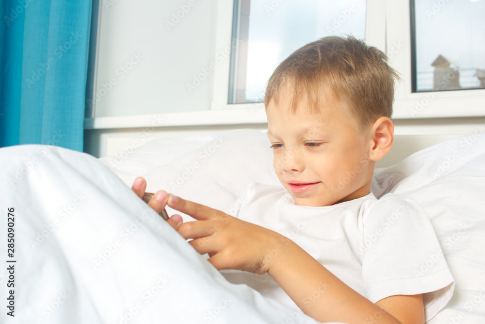 A six-year-old boy is resting lying in bed and holding a smartphone in his hands. A child with a gadget at home. A six-year-old boy is resting lying in bed and holding a smartphone in his hands. A child with a gadget at home.