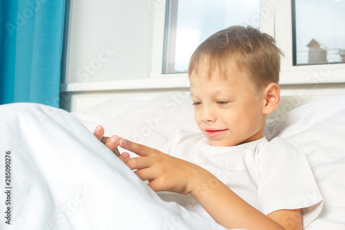 A six-year-old boy is resting lying in bed  and  holding a smartphone in his hands. A child with a gadget at home.