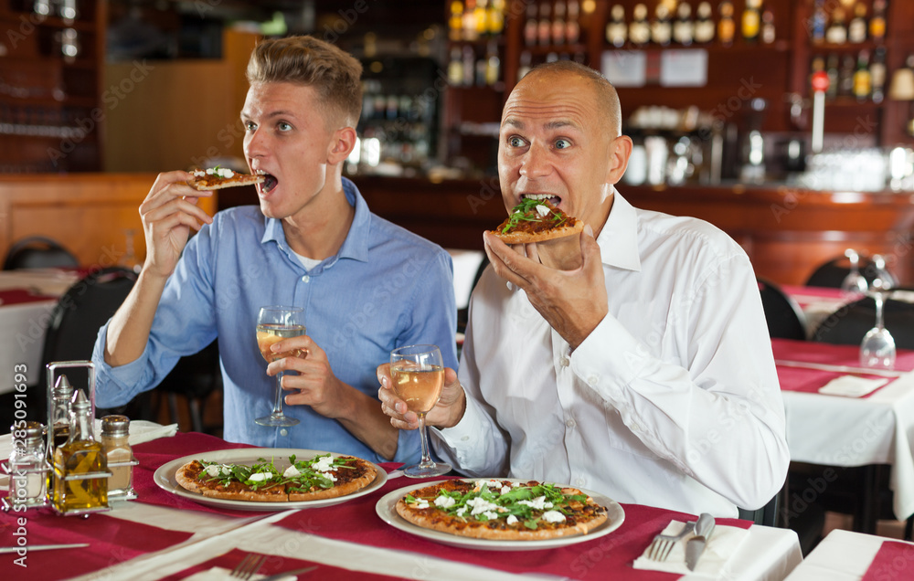 Two men watching sports while eating pizza in restaurant