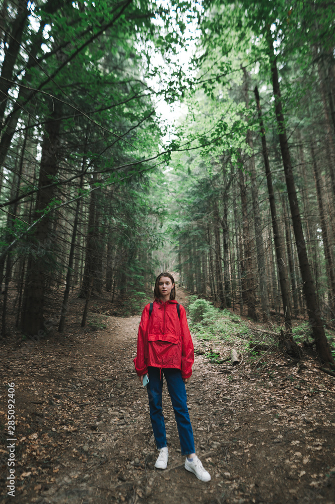 Young woman in a red jacket stands on a path in the forest, looks into the camera with a serious face. Portrait of hipster girl hiking through forest. Hiking mountains concept. Young woman in a red jacket stands on a path in the forest, looks into the camera with a serious face. Portrait of hipster girl hiking through forest. Hiking mountains concept.