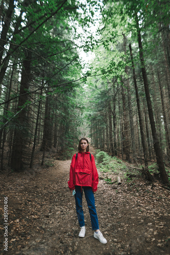 Young woman in a red jacket stands on a path in the forest, looks into the camera with a serious face. Portrait of hipster girl hiking through forest. Hiking mountains concept.