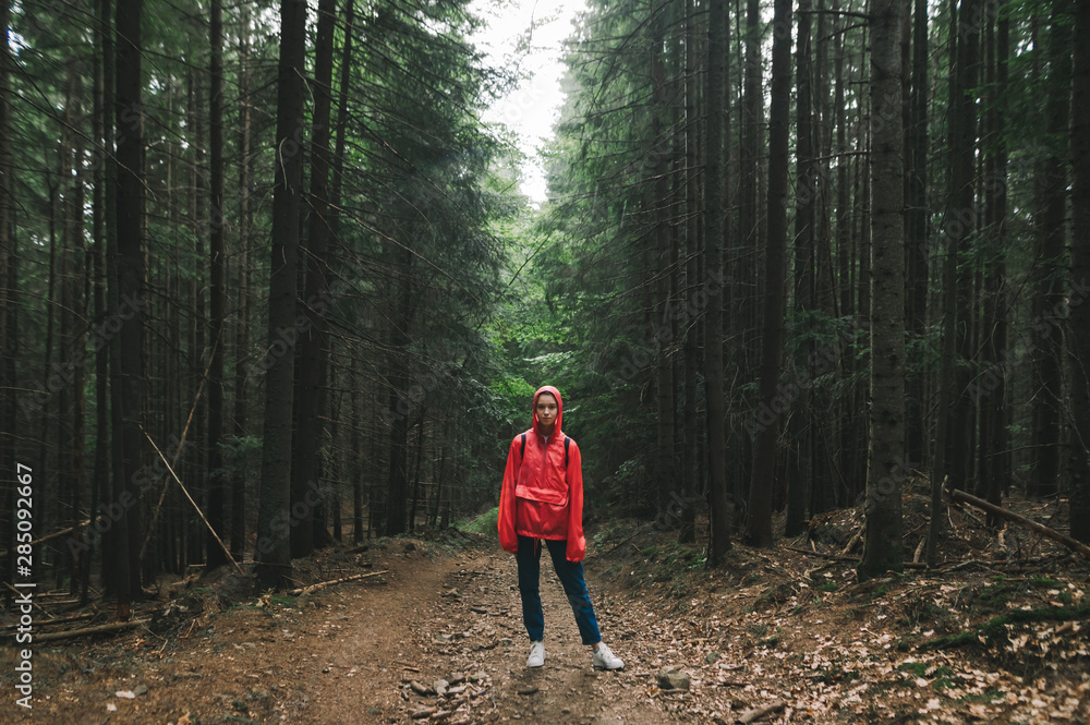 Obraz premium Hiker woman in a red raincoat stands on a trail in the mountains,looks in camera, girl on a mountain hike. Full length photo of tourist in red jacket standing in background of big old coniferous trees