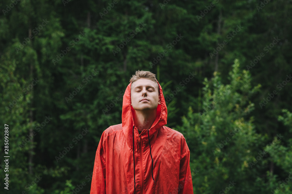 Handsome young man in a red raincoat stands outdoors in the rain with his eyes closed and enjoying the rain. Portrait of a young male hiker on a hike standing in the rain on a green background. Handsome young man in a red raincoat stands outdoors in the rain with his eyes closed and enjoying the rain. Portrait of a young male hiker on a hike standing in the rain on a green background.